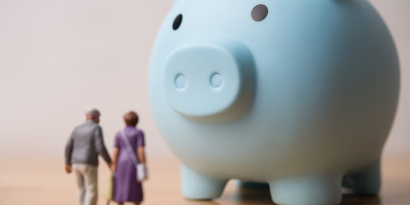Um casal de idosos em miniatura indo em direação de um cofrinho de porco para ilustrar a construção da Longevidade Financeira. 