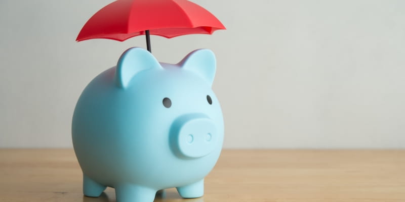 Um cofrinho de porco com um guarda-chuva representando a proteção financeira do capital, um dos pilares da Longevidade Financeira.