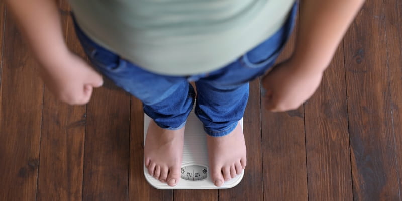 Menino com excesso de peso em pé sobre uma balança de chão dentro de casa, vista de cima. Imagem para ilustrar a matéria sobre obesidade infantil no Brasil.