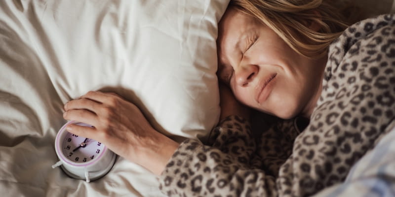 Um despertador em cima da almofada e uma mulher a dormir com uma expressão descontente por ter dificuldade ao acordar. Imagem para ilustrar a matéria sobre ritmo circadiano.