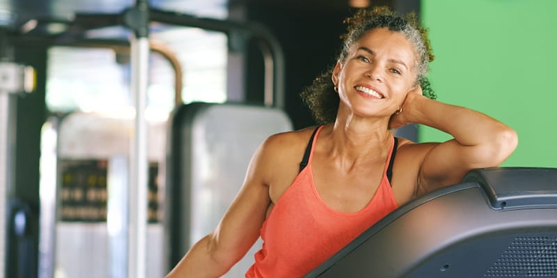 Uma mulher negra sorrindo apoiada em uma esteira de academia após se exercitar. Imagem para ilustrar a matéria sobre como envelhecer bem.