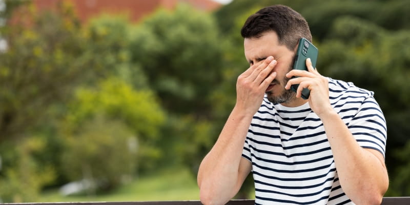 Um homem sentado em um banco de praça, com a mão no rosto, demonstrando desespero, enquanto fala ao telefone com o banco já que teve sua conta bloqueada pelo banco.