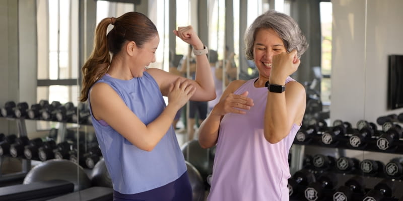 Uma mulher jovem e uma 60+, em uma academia, mostrando os músculos dos braços uma para a outra. As duas são asiáticas e sorriem. Imagem para ilustrar a matéria sobre estilo de vida para longevidade.