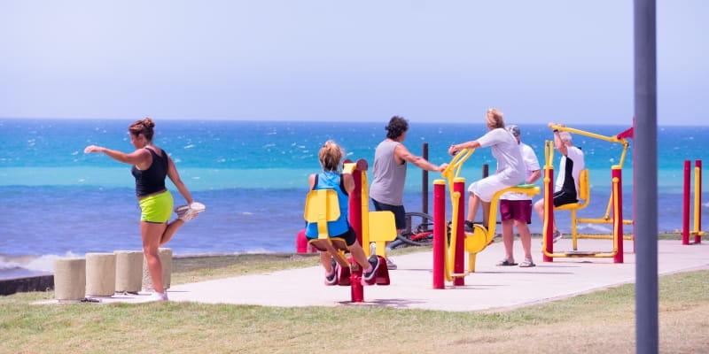 Idosos, em um litoral, fazendo atividade física em aparelhos de ginástica na praça, ilustrando matéria sobre longevidade saudável.
