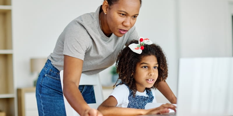 Uma mãe ensinando a filha. A mãe está em pé e a filha está sentada mexendo no computador portátil. Imagem para ilustrar a matéria sobre mulheres chefes de família.