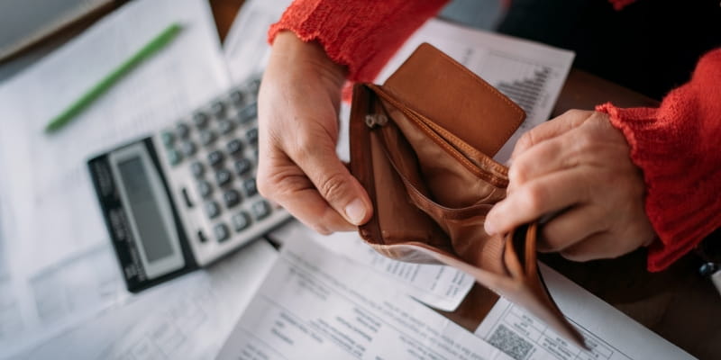 Mãos de uma mulher abrindo uma carteira vazia. Ao fundo, estão uma calculadora e contas para pagar. Imagem para ilustrar a matéria sobre renda insuficiente para despesas.