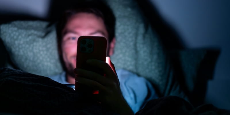 Um homem vendo conteúdo no celular de noite, enquanto está deitado na cama.
