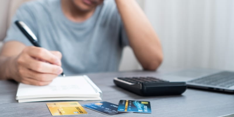 Um homem fazendo anotações em um caderno e com a mão na cabeça, demonstrando sua ansiedade financeira. Em sua frente, além do caderno, está uma calculadora e cartões de crédito.