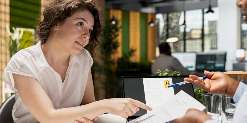 Uma mulher, em uma entrevista de emprego, enquanto o recrutador analisa seu currículo, negociando entre ser CLT ou PJ.