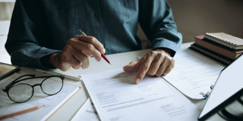Mãos de um homem sobre papéis a serem assinados, que estão sobre uma messa. Acima de um dos cadernos, está um óculos.