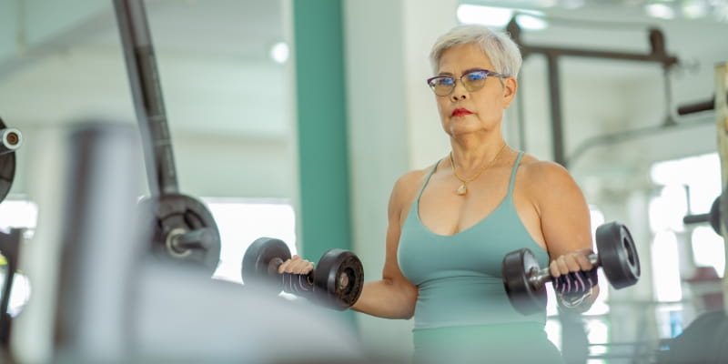 Uma mulher asiática e madura praticando musculação para envelhecer bem em uma academia. Ela está séria e levantando pesos.