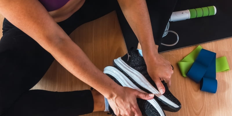 Uma mulher jovem, sentada no chão, com roupas de atividade física. Ao seu lado, estão elásticos de exercício, mostrando que ela faz atividade em casa.