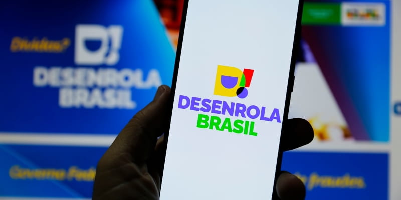 Uma pessoa segurando um celular com a logo do programa Desenrola Brasil, ilustrando a matéria que visa explicar quem pode participar do novo Desenrola Brasil.