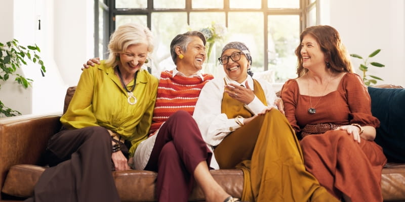 Um grupo de 4 mulheres maduras sem reunião em casa, sentadas no sofá e sorrindo. Imagem para ilustrar a matéria sobre amizades na maturidade.