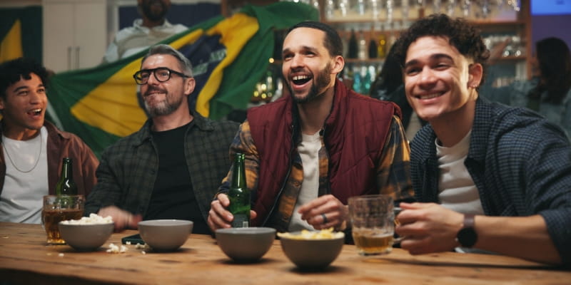 Um grupo de homens assistindo ao jogo do Brasil em um bar, comendo petiscos e bebendo cerveja. Imagem para ilustrar a matéria sobre como cuidar da imunidade durante a Copa do Mundo.
