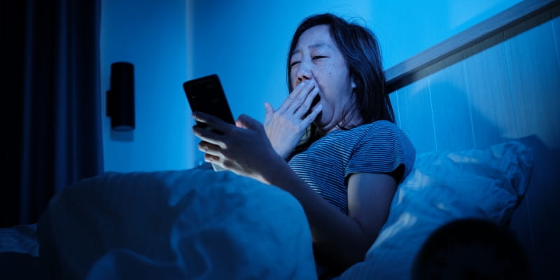Uma mulher asiática, deitada na cama, bocejando e mexendo no celular antes de dormir, mostrando que ela não faz as práticas ideiais de higiene do sono.
