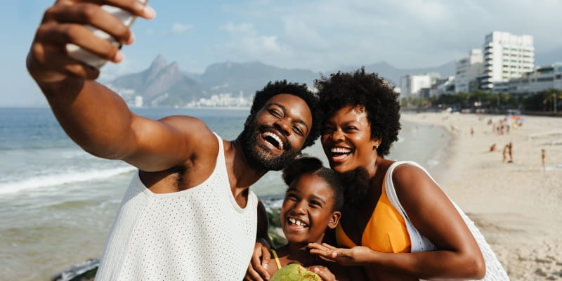 Uma alegre família brasileira desfruta de um dia ensolarado em uma praia no Rio de Janeiro, capturando o momento com uma selfie. Imagem para ilustrar a matéria sobre o Índice de Desenvolvimento Urbano para Longevidade.