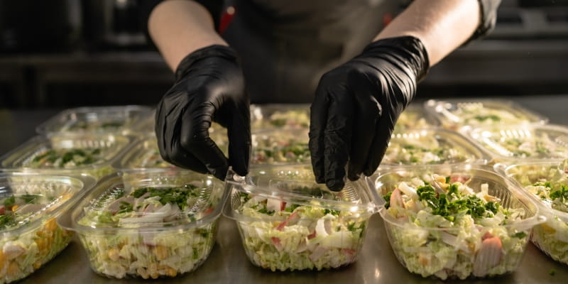 Uma profissional do mercado de marmitas montando potes de saladas enquanto usa luvas.
