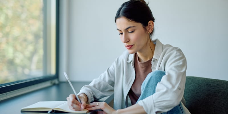 Uma jovem mulher com foco em escrever anotações em caderno aberto, estudando ou registrando sua mudança de personalidade por uma janela em luz natural.