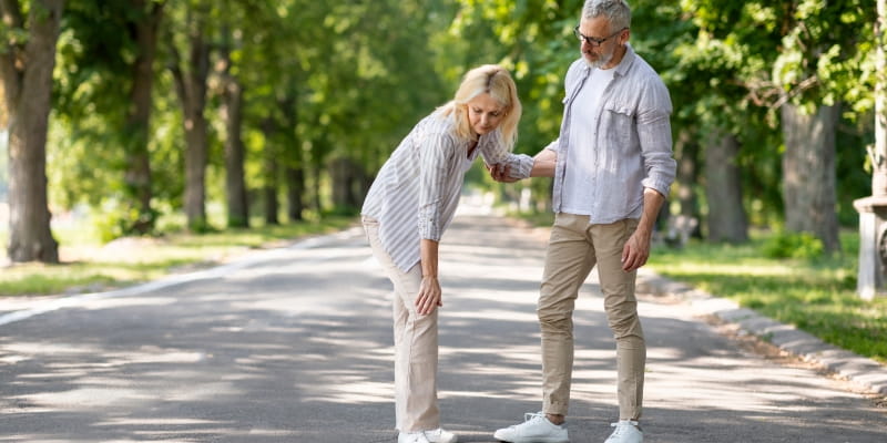Um casal passeando em um parque de área verde. A mulher se abaixa, demonstrando dor nas pernas enquanto o homem a segura. A imagem ilustrar a matéria sobre a perda de força muscular.
