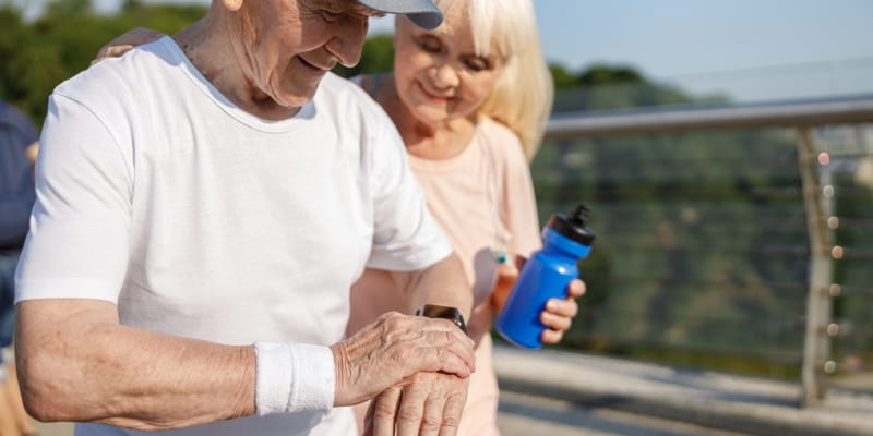 Homem idoso de boné monitora a frequência cardíaca com um smartwatch durante o treino com a esposa. Imagem para ilustrar a matéria sobre reduzir o risco cardiovascular.