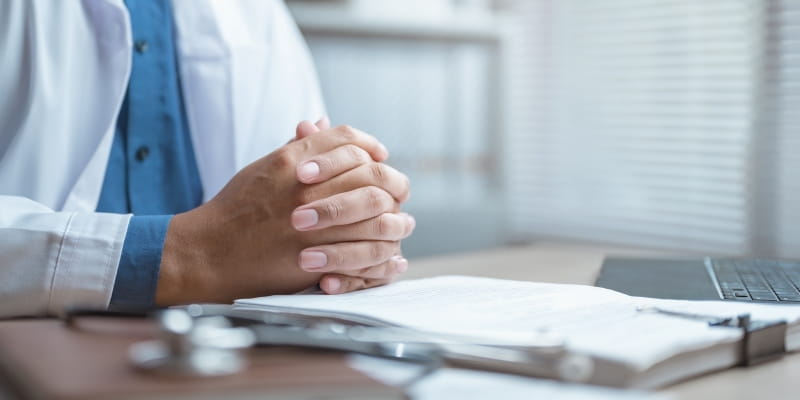Mãos de um médico sobre uma mesa, com um computador e papéis da pesquisa sobre câncer em jovens.