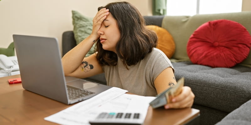 Jovem mulher, em frente ao computador, segurando notas de dinheiro e tentando fazer um controle financeiro pessoal com uma calculadora. Ela está com a mão no rosto, demonstrando frustração.