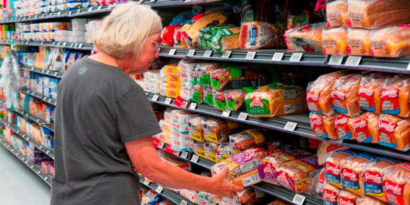 Uma mulher idosa fazendo compras de supermercado e escolhendo pão de forma na prateleira. Imagem para ilustrar a matéria sobre desigualdade em saúde.