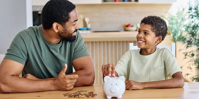 Um pai ensinando sobre finanças para o filho enquanto o adolescente coloca moedas em um cofrinho. Imagem para ilustrar a matéria sobre educação financeira para jovens.
