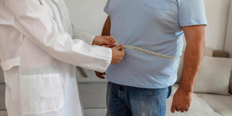 Um homem sendo avaliado por um médico enquanto o especialista passa uma fita métrica sobre sua região abdominal para verificar seu peso e circunferência.
