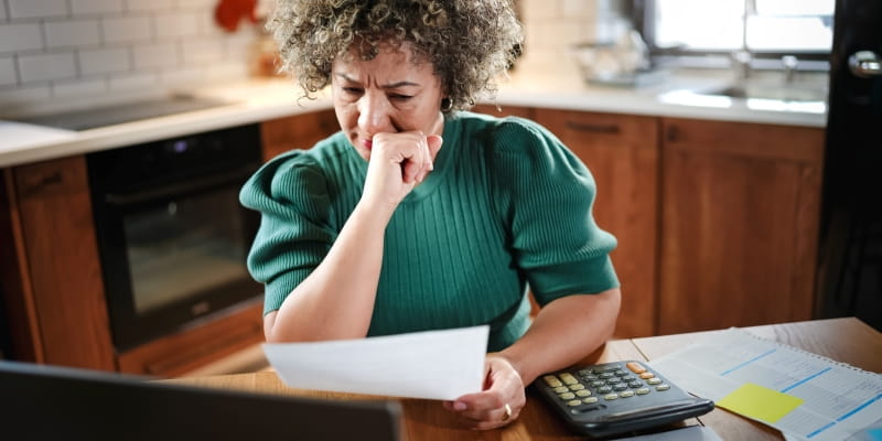 Uma mulher madura, negra, com cabelos grisalhos pcupada revisando papelada em sua mesa na cozinha. Ela segura um documento olhando atentamente para ele e na mesa estão um laptop, uma calculadora e papéis. Imagem para ilustrar a matéria sobre independência financeira feminina.