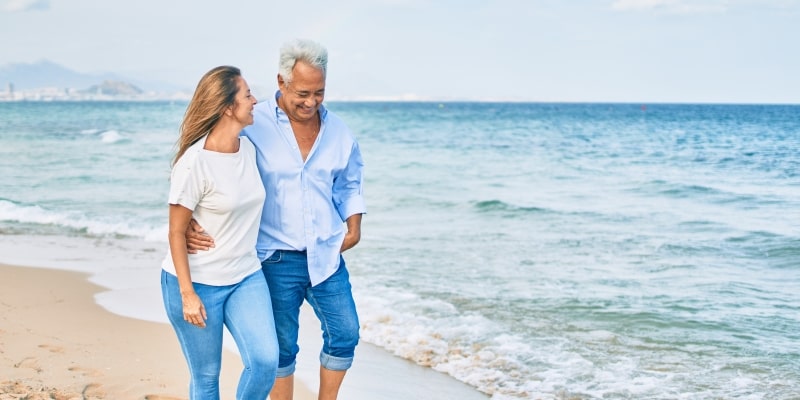 Um casal de idosos andando na praia enquanto se olham e sorriem. Imagem para ilustrar a matéria sobre melhores países para se aposentar.