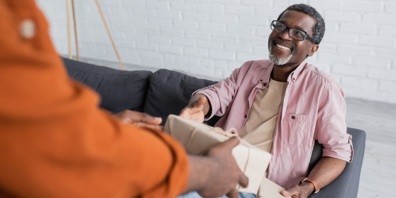 Um homem negro, com cerca de 50 anos, sentado em um sofá, sorrindo e com a mão estendida, recebendo um presente de Dia dos Pais.